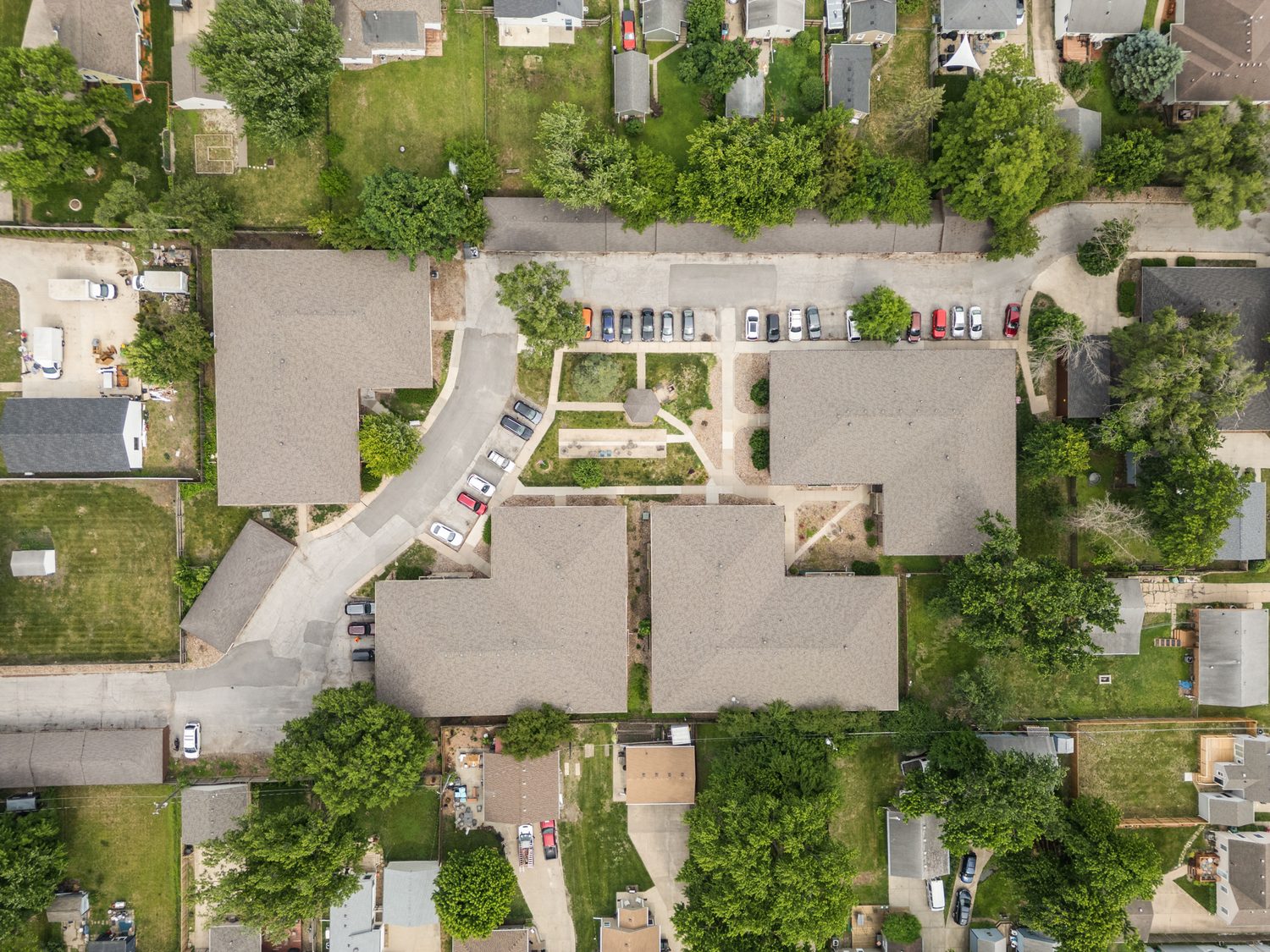 Aerial view straight down on Trailside, four brick apartment buildings with grey roofs around a central lawn and gazebo, a drive with parked cars running between them, and mature trees on every side.