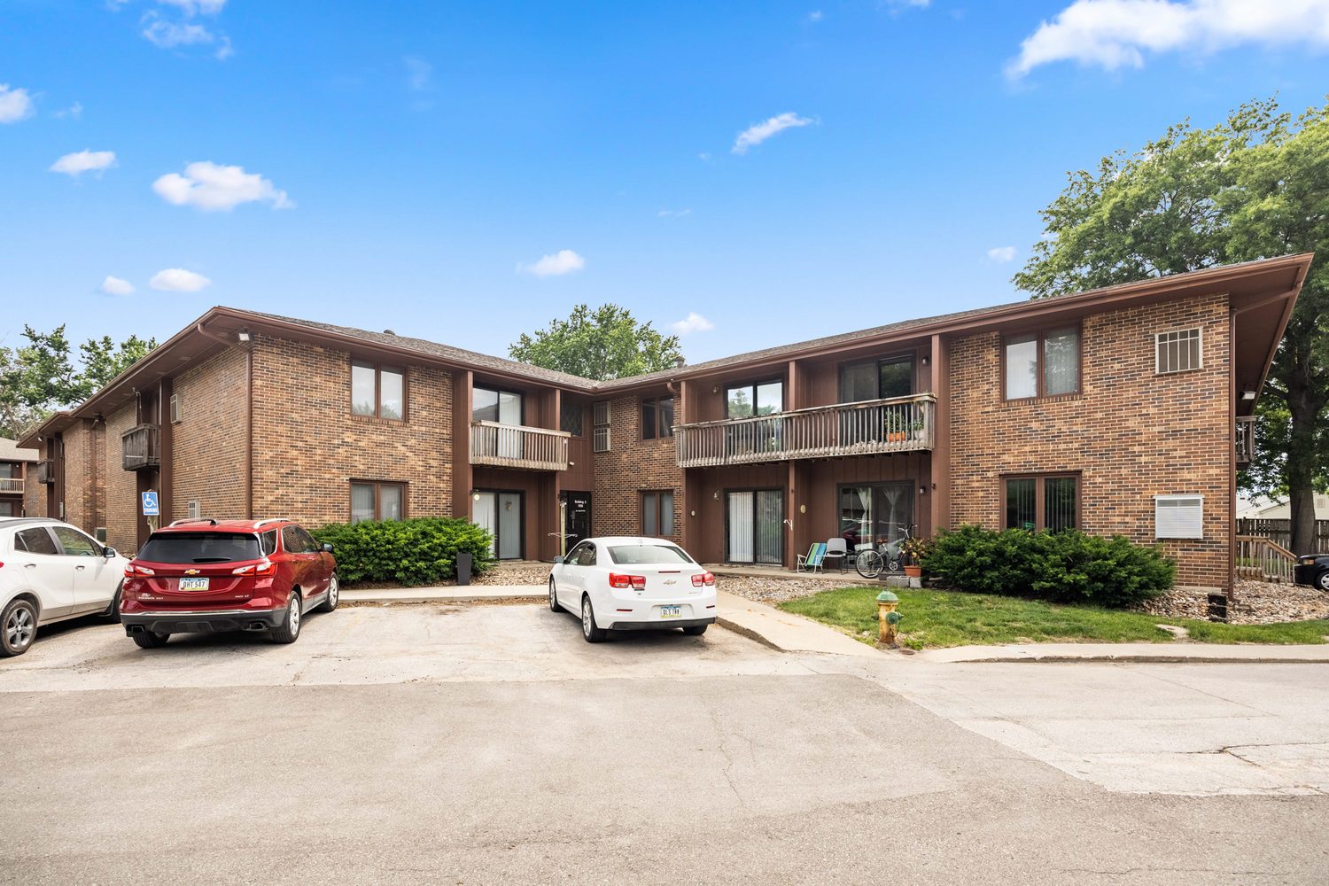 Two Trailside buildings from the drive in summer, two-story orange-brown brick with dark wood trim, second-floor balconies and ground-floor patios, and cars parked along the drive.