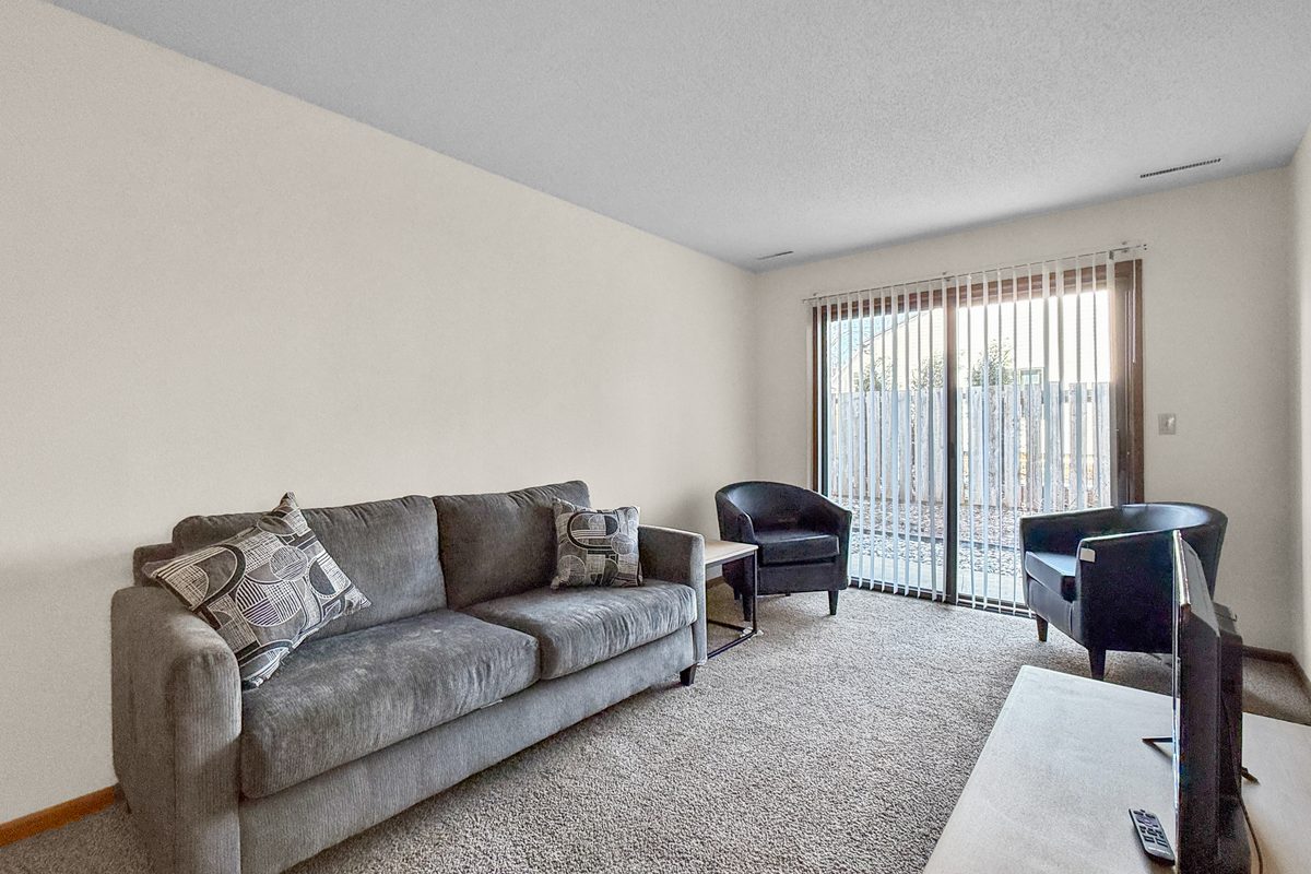 A furnished living room with grey carpet, a grey sofa and two black chairs, and a sliding glass door with vertical blinds out to the patio.