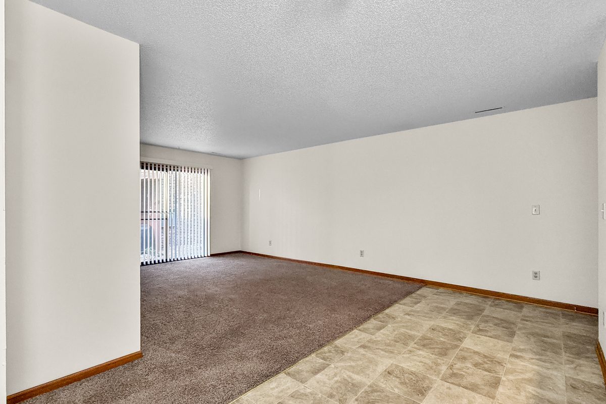 The living room of a two bedroom apartment, carpeted, open to the kitchen with oak cabinets and a white refrigerator.