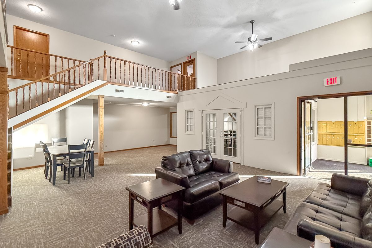 A two-story building lounge with a wood staircase, grey couches, a dining table with chairs and a bookshelf.