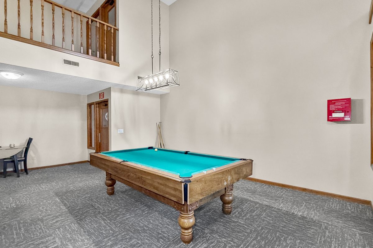 A pool table with a green felt top in a building lounge, with a ceiling light above it and a window to the drive.