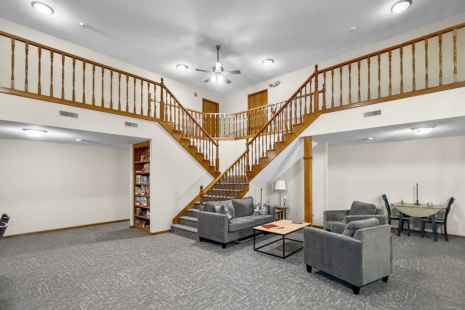 A two-story resident lounge at Trailside with a wood staircase and railing, grey couches around a coffee table, a dining table with chairs, and a bookshelf under the stairs.