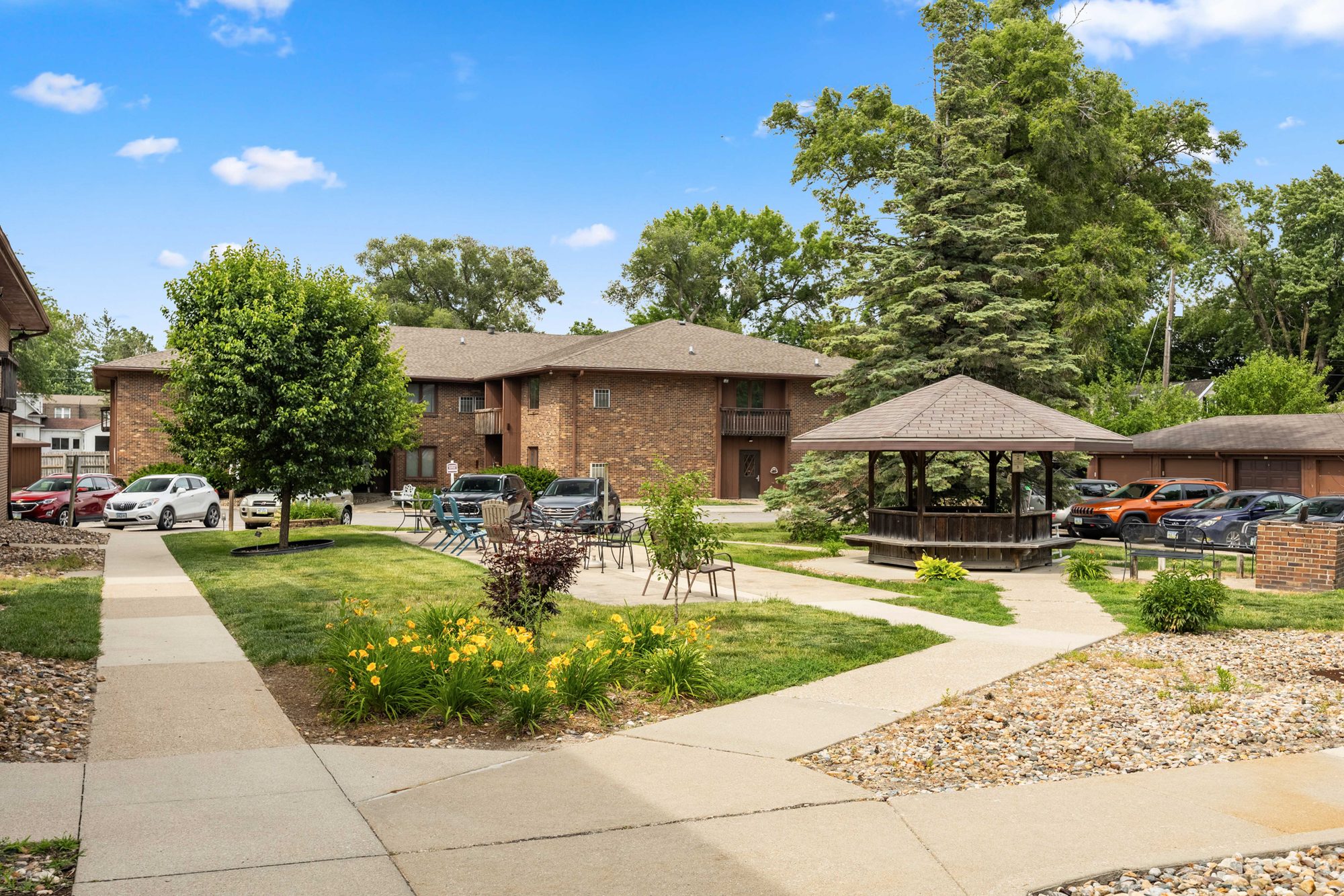 The central green at Trailside Apartments in summer, a wood gazebo on a lawn with flower beds and tall mature trees, with the two-story brick apartment buildings behind it.